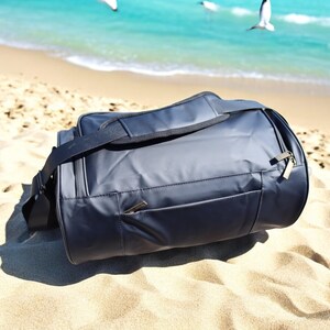 May include: A black duffel bag with a zippered pocket on the front. The bag is sitting on a sandy beach.