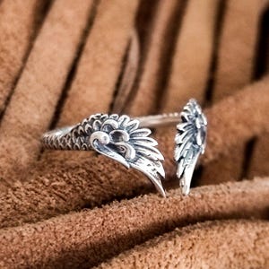 Op de afbeelding: Een zilverkleurige ring met een gedetailleerd vleugelontwerp. De ring heeft twee gestileerde vleugels die zich om de vinger wikkelen. De ring staat tegen een bruine, getextureerde achtergrond. De ring is een sieraad.