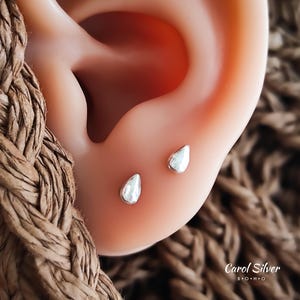 May include: Close-up of a person's ear with two small silver teardrop-shaped earrings. The earrings are simple and delicate, and they are a popular choice for everyday wear.