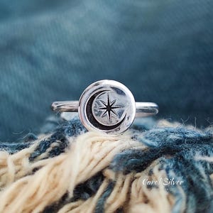 Op de afbeelding: Zilveren ring met een cirkelvormig ontwerp met een maan en een ster. De ring staat tegen een blauw en wit getextureerde achtergrond. De ring is een sieraad met een hemels thema.