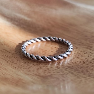 May include: A silver ring featuring a twisted rope design. The ring is circular and rests on a wooden surface. The ring's texture is visible, with each twist of the rope clearly defined. The ring is a piece of jewellery.