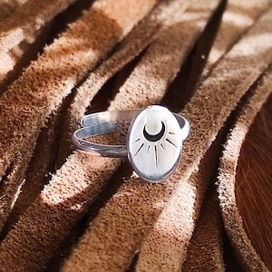 Może przedstawiać: Srebrny pierścionek z owalną tarczą z motywem półksiężyca i promieni słonecznych. Pierścionek jest umieszczony na brązowym skórzanym tle, prezentując szczegóły i kunszt biżuterii. Pierścionek ma prosty, podwójny wzór obrączki.