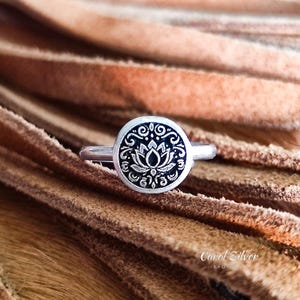 May include: A silver ring with a circular face featuring a lotus flower design. The ring's face has a black background with a white lotus flower and decorative patterns. The ring is set against a brown, textured background.