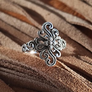 Puede incluir: Un anillo plateado con un diseño floral ornamental y arremolinado. El anillo presenta un grupo central de pequeñas esferas rodeadas de intrincados adornos y florituras. El anillo se muestra sobre una superficie marrón.