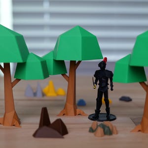 Könnte beinhalten: Eine Miniatur-Waldszene mit grünen geometrischen Bäumen und einer schwarzen Figur mit rotem Hut. Braune Baumstämme und verschiedene farbige geometrische Formen sind in der Szene verstreut und schaffen eine spielerische, fantasievolle Umgebung.