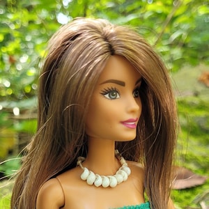 May include: A doll with long brown hair wearing a green tube top, matching skirt, and a shell necklace. The doll is posed outdoors against a backdrop of green foliage and moss-covered rocks. The skirt has a slit.
