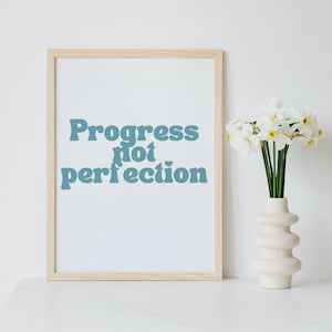 May include: A framed print with the text "Progress not perfection" in blue lettering on a white background. The print is sitting on a white surface next to a vase of white daffodils.