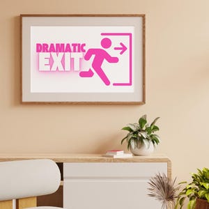 May include: A framed print with the words "DRAMATIC EXIT" in pink neon lettering, accompanied by a pink figure running towards an exit sign. The artwork is displayed on a light-colored wall above a white cabinet with plants.