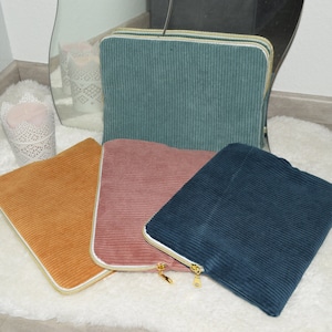 Peut inclure: Quatre étuis pour ordinateur portable en velours côtelé de couleurs différentes avec des bordures blanches et des fermetures éclair dorées. Les étuis sont disposés sur une surface blanche et moelleuse.