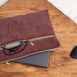 Op de afbeelding: Een mauve corduroy laptophoes met een gouden rits. De hoes is open en toont een laptop en zwarte koptelefoon. Een zwarte computermuis ligt op het houten oppervlak.