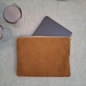 May include: Mustard-colored corduroy zippered pouch with a gold zipper, holding a gray tablet. The pouch is rectangular and has vertical ribbed texture. Two pink candles in white lace holders are in the background.