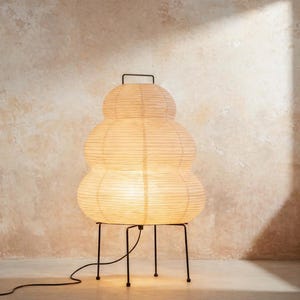 May include: A unique, stacked paper lantern lamp with a warm, inviting glow. The lamp has a cream-colored, layered shade and sits on a black metal frame. The lamp's cord extends to the left, suggesting a power source.