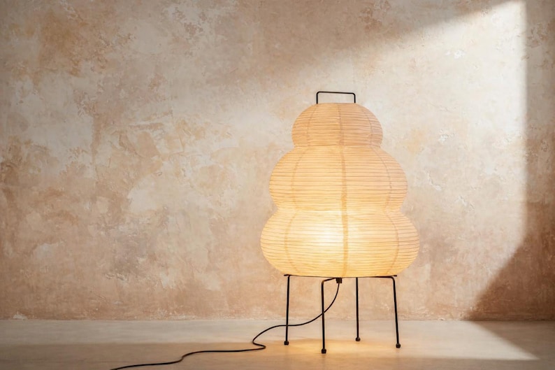 May include: A unique, stacked paper lantern lamp with a warm, inviting glow. The lamp has a cream-colored, layered shade and sits on a black metal frame. The lamp's cord extends to the left, suggesting a power source.