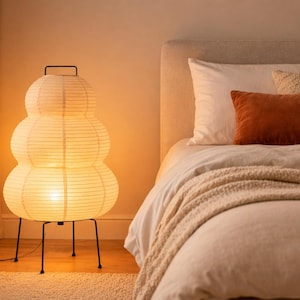 Puede incluir: Una l&aacute;mpara de pie de papel con un dise&ntilde;o apilado y redondeado, iluminada desde dentro. La l&aacute;mpara tiene una estructura y un asa de metal negro. La luz c&aacute;lida ilumina un dormitorio con una cama, almohadas y una manta. La habitaci&oacute;n tiene una paleta de colores neutros.