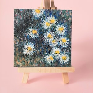 May include: A small painting on a wooden easel depicts a cluster of white daisies with yellow centers against a dark blue and green background.
