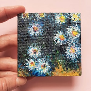 May include: A small painting on canvas featuring a cluster of white and blue flowers with yellow centers, set against a dark green background.