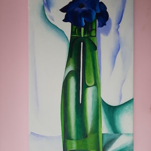 May include: A painting of a single blue flower in a green glass vase. The vase is on a white surface with a blue and white background. The painting is signed "Georgia O'Keeffe".