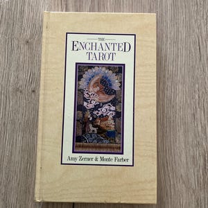 May include: A hardback book titled "The Enchanted Tarot" by Amy Zerner & Monte Farber. The cover features a colourful illustration of a crescent moon, stars, and a building. The book has a light tan cover.