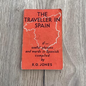May include: A vintage travel guide titled "The Traveller in Spain" with a red cover. The book features a white outline of Spain and text that reads "useful phrases and words in Spanish". Compiled by R.O. JONES.