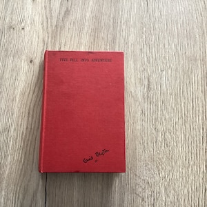 May include: A red hardback book with the title "Five Fall Into Adventure" and the author's name "Enid Blyton" written on the cover.