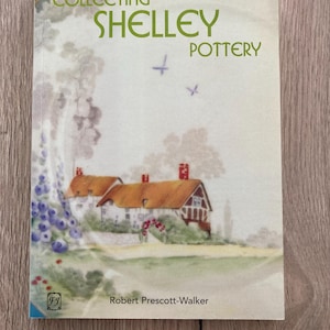 May include: A softcover book titled Collecting Shelley Pottery by Robert Prescott-Walker. The cover features a watercolour illustration of a cottage with a thatched roof, purple flowers, and two birds flying in a pale sky against a white background.