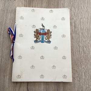 May include: A vintage booklet with a white cover featuring a coat of arms and crown pattern. A ribbon in red, white, and blue is tied around the booklet. The booklet is on a wooden surface.