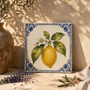 Pode incluir: Um azulejo de cerâmica quadrado com uma ilustração central de um limão amarelo brilhante com folhas verdes e uma flor branca. O azulejo é emoldurado por uma borda floral azul decorativa sobre um fundo creme, com design botânico.