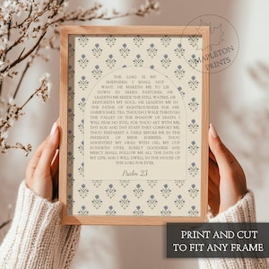 May include: A framed print featuring Psalm 23 text centered within an arched design. The background displays a repeating pattern of small blue floral motifs on a cream-colored surface. The frame is made of light-toned wood with a simple profile.
