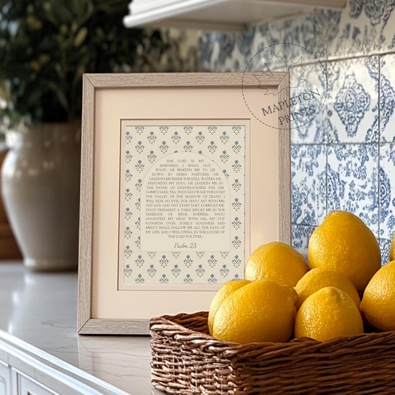 May include: A framed print featuring the text of Psalm 23 centered within an arched design, surrounded by a repeating blue floral pattern on a cream background. The frame is light wood, and the print is displayed in a kitchen setting.