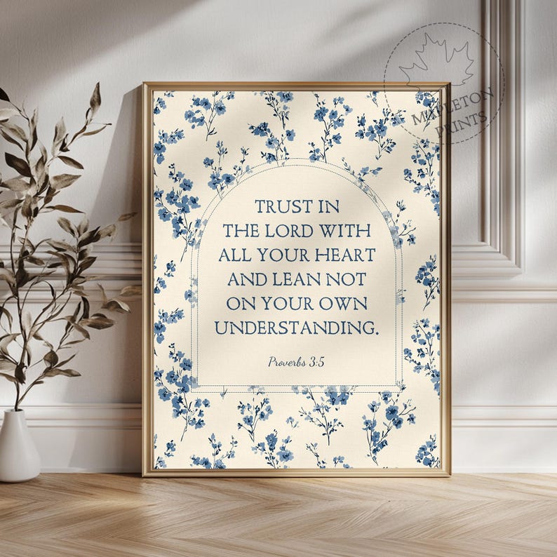 May include: A framed print featuring the text "Trust in the Lord with all your heart and lean not on your own understanding. Proverbs 3:5." The background is a cream color with blue floral accents, and the frame is gold.