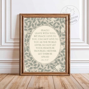 May include: A framed print featuring the text "PEACE I LEAVE WITH YOU; MY PEACE I GIVE TO YOU..." within an oval, encircled by a floral pattern. The frame is brown, and the background is a light beige with green and white floral details. The print is on a wooden surface.