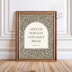 Op de afbeelding: Een ingelijste print met de tekst "GIVE US THIS DAY OUR DAILY BREAD" uit Mattheüs 6:11. De tekst staat in een witte boogvorm tegen een donkergroen bloemmotief. De lijst heeft een warme bruine kleur.
