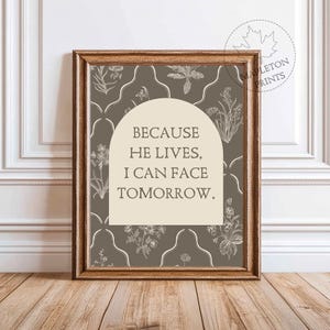 Op de afbeelding: Een ingelijst drukwerk met een bruine achtergrond en een wit bloemenpatroon. De print bevat een witte boog met de tekst "Because He Lives, I Can Face Tomorrow."