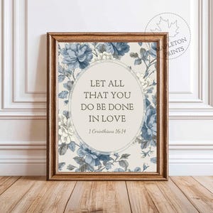 May include: Framed print with a floral design in shades of blue and white. The print features the text "LET ALL THAT YOU DO BE DONE IN LOVE" and "1 Corinthians 16:14" in a central oval. The frame is a warm brown color.
