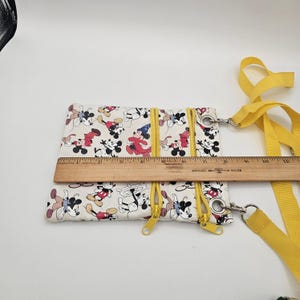 May include: A white crossbody bag featuring a repeating Mickey Mouse print with yellow zips and a yellow strap. A wooden ruler placed across the bag shows a width of approximately 20 cm. The bag has two front zipped compartments.