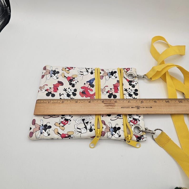 May include: A white crossbody bag featuring a repeating Mickey Mouse print with yellow zips and a yellow strap. A wooden ruler placed across the bag shows a width of approximately 20 cm. The bag has two front zipped compartments.