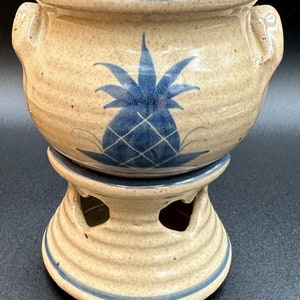 May include: A ceramic pot with a blue pineapple design and a matching stand. The pot has two handles and the stand has three open spaces.
