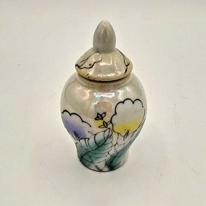 May include: A small white ceramic jar with a lid, decorated with a floral design in shades of purple, yellow, and green.