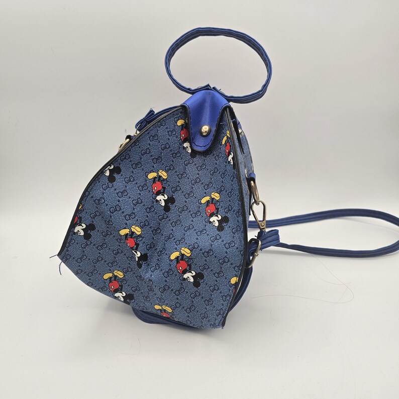May include: A triangular-shaped handbag featuring a blue denim-like fabric with a repeating pattern of Mickey Mouse figures and the letters GG. The bag includes a circular top handle, blue trim, a gold-tone metal stud, and a detachable shoulder strap.