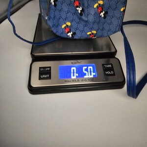 May include: A blue bag with a repeating Mickey Mouse pattern and a monogram print, resting on a digital scale that displays a weight of 141.7 grams. The bag features thin blue straps and a structured base visible on the scale platform.