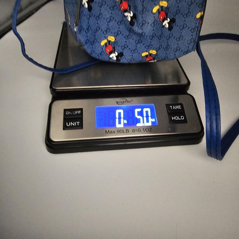 May include: A blue bag with a repeating Mickey Mouse pattern and a monogram print, resting on a digital scale that displays a weight of 141.7 grams. The bag features thin blue straps and a structured base visible on the scale platform.