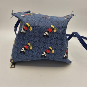 May include: A blue crossbody bag featuring a repeating pattern of Mickey Mouse characters and a dark blue monogram print. The bag has a structured, boxy shape with a top zipper closure, a gold-toned metal zipper pull, and a thin blue shoulder strap.