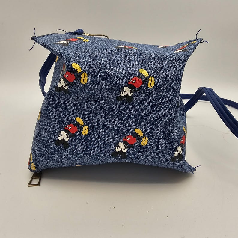 May include: A blue crossbody bag featuring a repeating pattern of Mickey Mouse characters and a dark blue monogram print. The bag has a structured, boxy shape with a top zipper closure, a gold-toned metal zipper pull, and a thin blue shoulder strap.