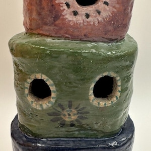 May include: A three-tiered ceramic planter with a unique design. The top tier is pink with a black and white flower design and a hole in the center. The middle tier is green with two holes and a black and white flower design. The bottom tier is blue with a single hole in the center.