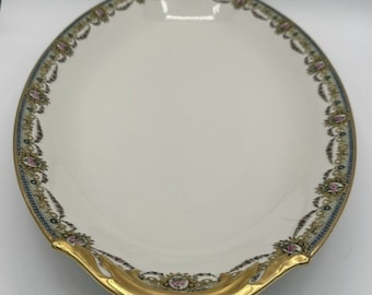 May include: An oval porcelain serving platter with a white center and a decorative border featuring gold trim, blue accents, and small pink floral motifs. The rim has a scalloped edge with prominent gold-painted sections at the top and bottom.