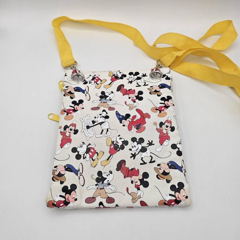 May include: A rectangular crossbody bag featuring an off-white fabric base with a repeating pattern of Mickey Mouse in various outfits and poses. The bag includes a bright yellow adjustable strap and yellow zip pulls attached to metal hardware.