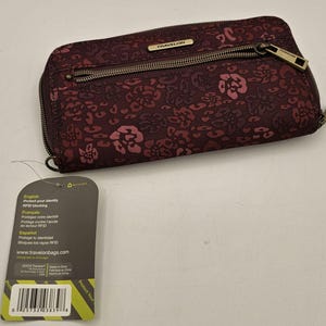 May include: A dark burgundy Travelon wallet featuring a repeating floral and animal print pattern in lighter pink tones. The wallet includes a front zippered pocket with a metallic zipper pull and a metal brand plaque. A product tag is attached.
