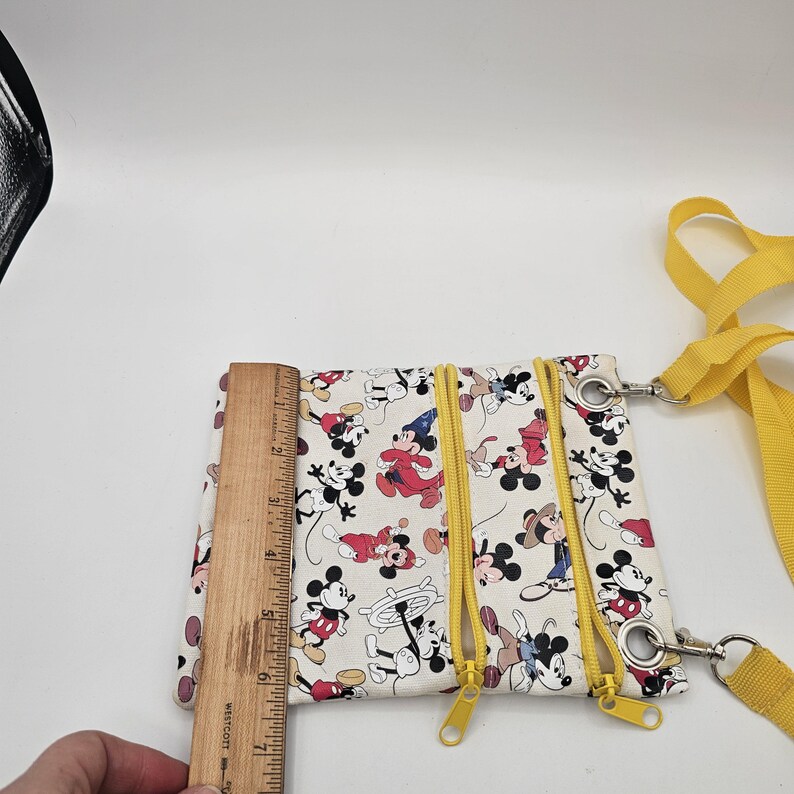 May include: A cream-coloured crossbody bag featuring a repeating Mickey Mouse print in various outfits, including a wizard and sailor. The bag has two yellow zippered compartments and a yellow strap. A wooden ruler shows a height of 17.8 cm.