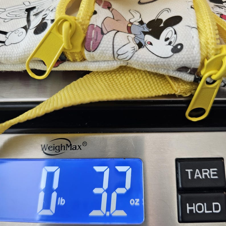 May include: A white canvas pouch featuring a Mickey Mouse print with yellow zips and a yellow strap. The pouch is resting on a digital scale that displays a weight of 90.7 grams, clearly visible on the blue backlit screen of the device.