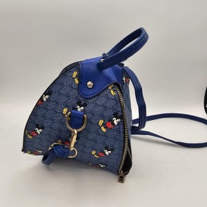 May include: A blue triangular handbag featuring a repeating pattern of Mickey Mouse figures and a small logo print. The bag includes a solid blue top handle, a silver-toned metal zip closure, and a detachable blue shoulder strap with gold-toned hardware.
